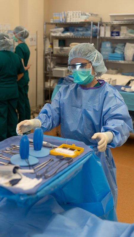 photo of member of Surgical Technology program dressed in gown, cap, gloves and mask standing behind a table with tools on it.