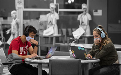 two students sitting at a table in the library with masks on working on laptops