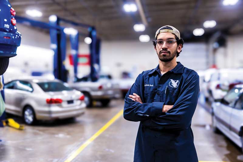 Auto Tech student in the work area on campus with cars on lifts int he background