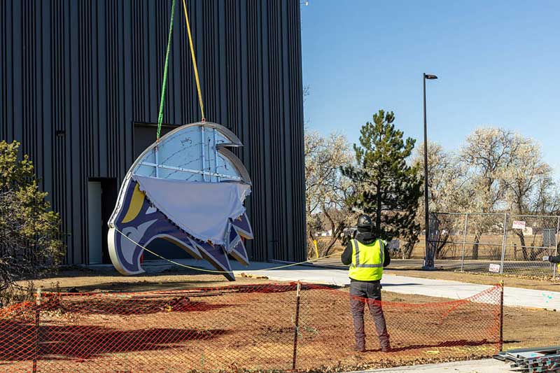 A photo of the large athletics logo sign being installed by crane and lifted by a strap off the ground while a worker watches