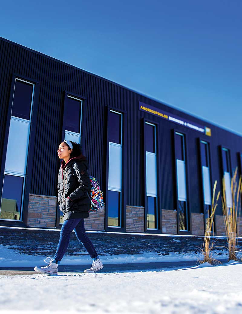a Student walking across campus in the winter with the Business building in the background