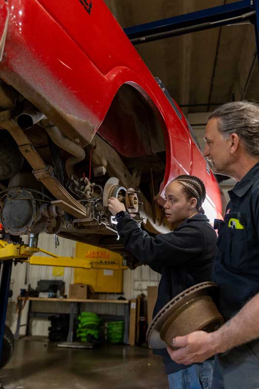 Jaya Brown standing beside a car in the Auto Tech bay with an instructor working on the wheel