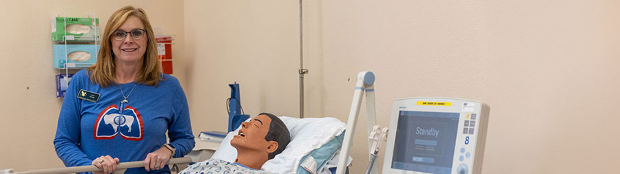 Lori Arnold standing in a simulation space with a simulation mannequin in a hospital bed with equipment.