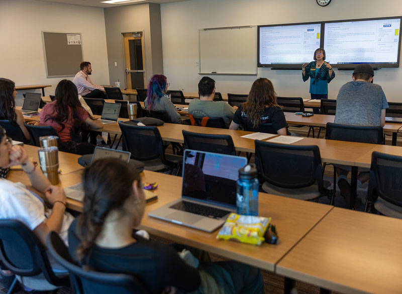 photo of students in a classroom with laptops and an instructor in the front of the room with information on the board behind her.