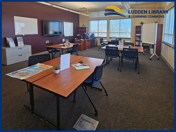 Several table groupings and one large desk. Wall screen on left side. Ludden library & Learning commons logo in upper right corner