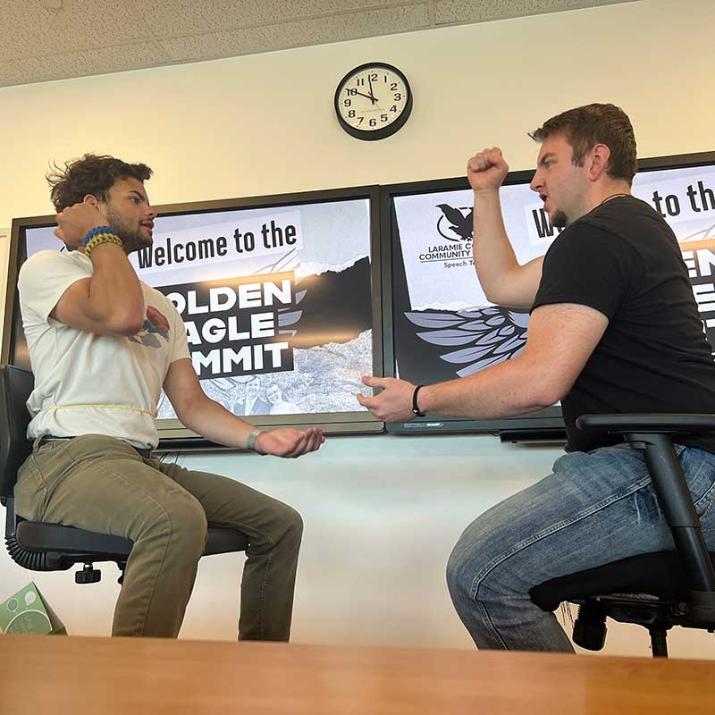 Two students sitting in chairs in a classroon doing rock, paper, scissors