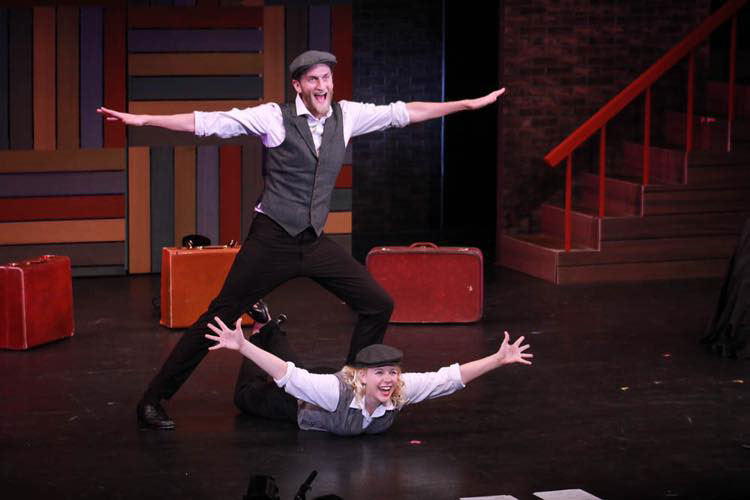 Photo of two actors on stage wearing caps and vestswith suitcases behind them. One actor is laying on the ground with arms outstretched. The other is standing over them with arms outstretched.