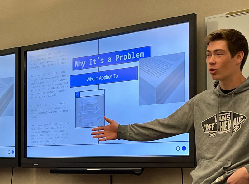 Photo of a student presenting in front of two screens with a PowerPoint presentation on display behind him.