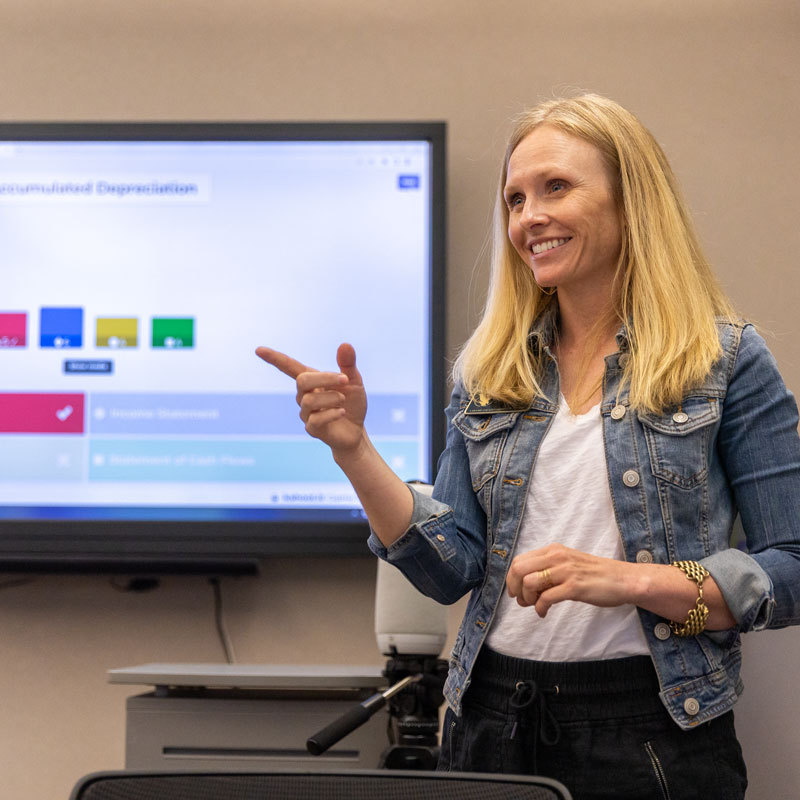 photo of a female instructor speaking to a class and pointing while standing front of a screen with a presentation on it.