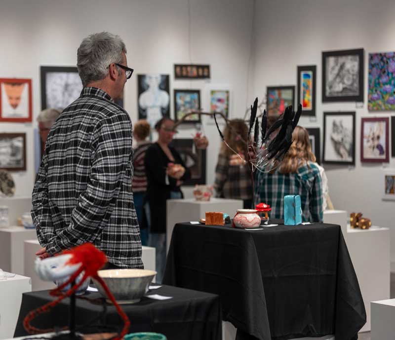 A person looking at the 3D artwork in the middle of the gallery during the 2025 LCSD1 High School art show. 2D work is on the walls in the background.