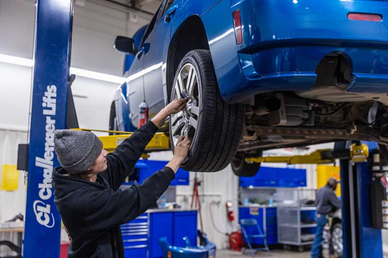 A student tightens lug nuts on the back tire of a car that is lifted in the repair area.