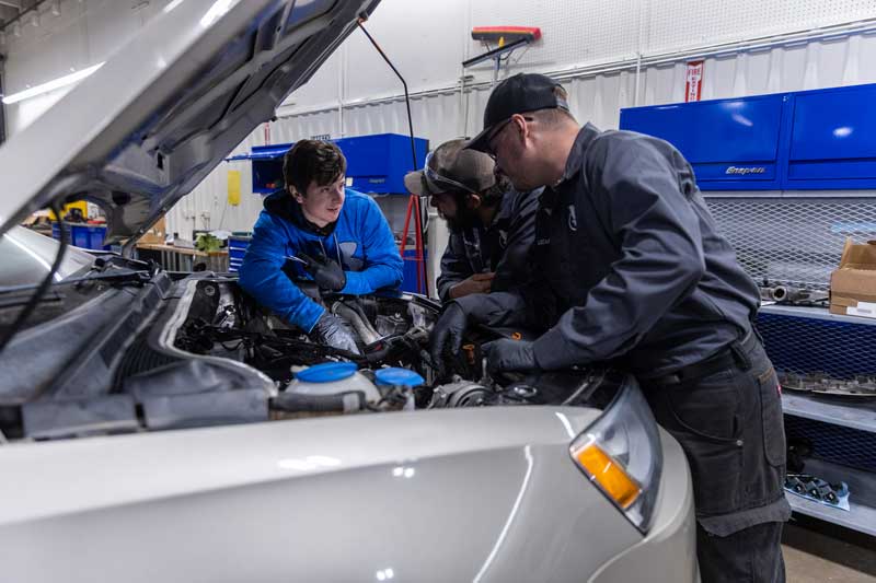 A student and two instructors are leaning into the engine area of a car with the hood up talking and pointing.