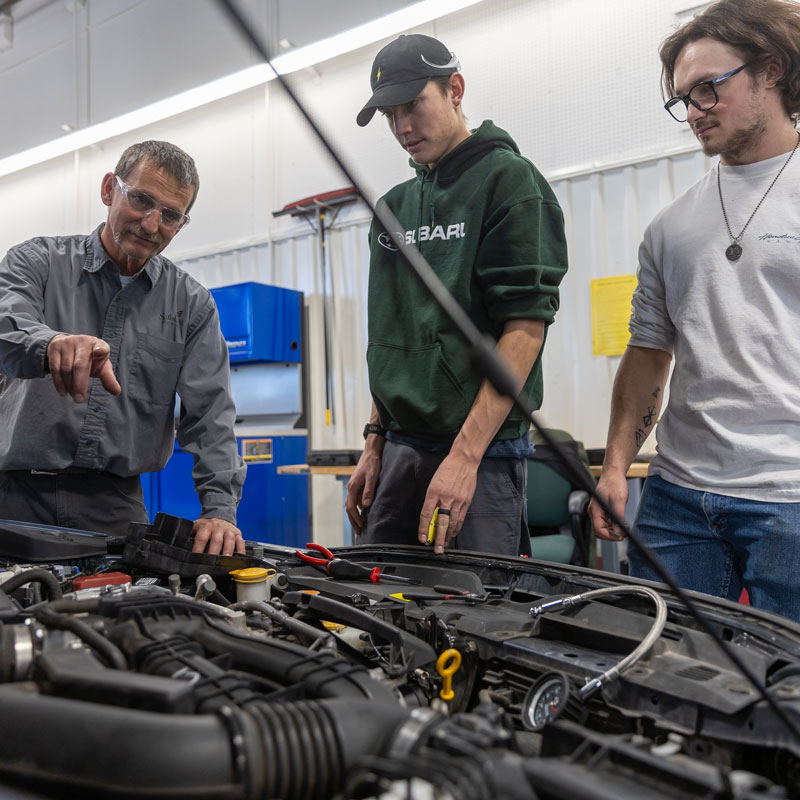 photo of faculty member and two students looking under the hood of a car. Faculty is pointing at something.