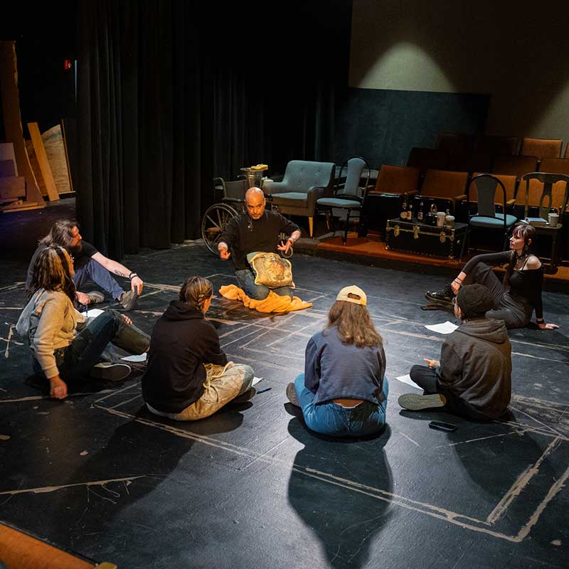 Students in acting class sitting on the floor in The Playhouse theatre with prop behind them as well as seats for the audience