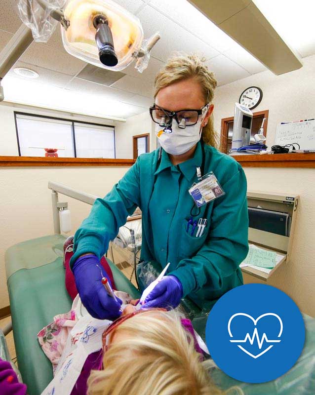 Dental hygiene student giving cleaning to child with an icon in the lower right corner for the Health Sciences and Wellness Pathway with a heart and heartbeat.