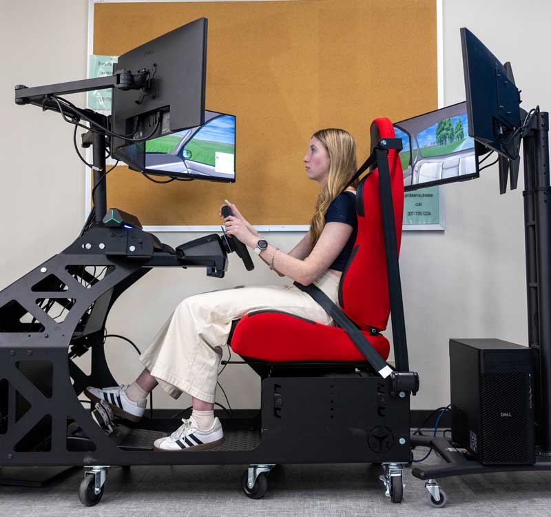 Side view of a student driver in the driving simulator showing the front screens, steering wheel, seat with seatbelt and rear screens