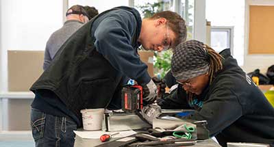 Two Metallica Scholars who are using a drill to attach materials on a table in class