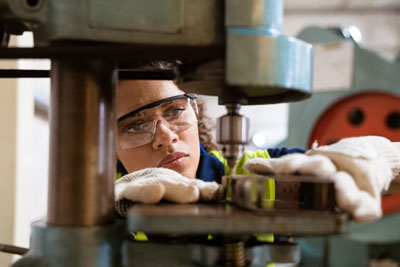 photo of a female worker drilling material on a drill press with safety glasses on
