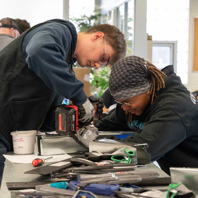 two students using a drill to assemble some metal pieces on a table